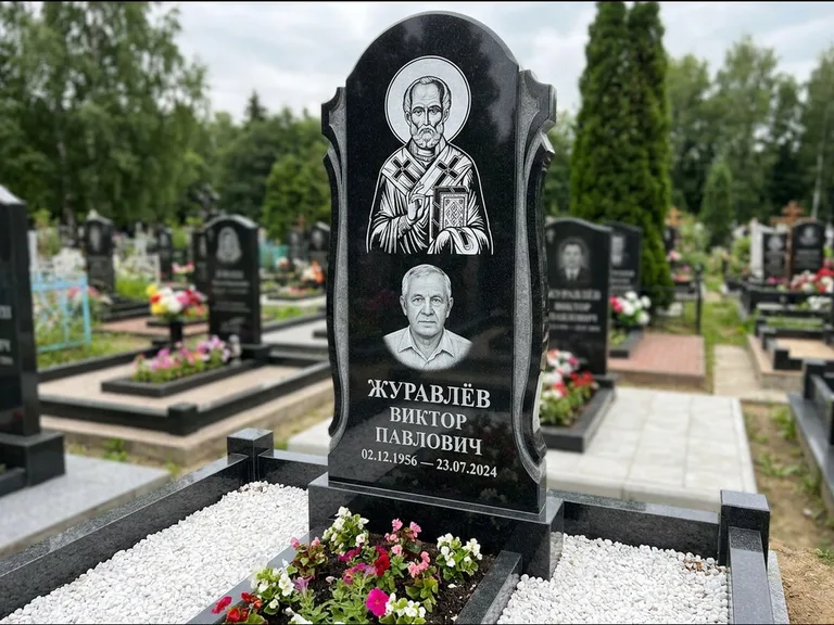 Памятник с гравировкой иконы: стела с образом в полный рост на обороте — гравировка образа Матроны Московской, низкие кусты спиреи в цвету, работа «Лебедева» в Москве